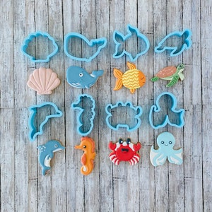 May include: A set of 14 blue plastic cookie cutters shaped like various sea creatures, including a whale, a seahorse, a starfish, a crab, an octopus, a seashell, and a dolphin. The cutters are arranged on a light wood background.