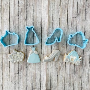 May include: A set of blue plastic cookie cutters in the shape of a crown, dress, shoe, carriage, and a dress with a crown. There are also two cookies decorated with frosting in the shape of a crown and a dress.