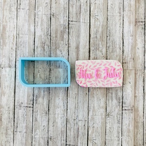 May include: Two cookie cutters, one light blue and one pink with a white background and a pink leaf pattern. The pink cutter has the text "Alex & Julie" in pink.