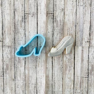 May include: A blue plastic cookie cutter shaped like a high-heeled shoe next to a silver-colored sugar cookie decorated to look like a high-heeled shoe.