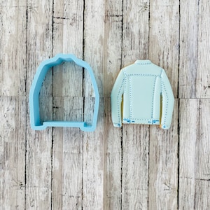 May include: A light blue cookie cutter shaped like a denim jacket, next to a decorated cookie in the shape of a denim jacket.