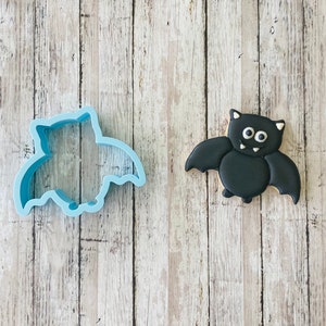 Puede incluir: Un cortador de galletas de murciélago de plástico azul y una galleta de azúcar negra decorada para parecer un murciélago. La galleta tiene ojos blancos y una sonrisa blanca.