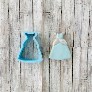 May include: A light blue cookie cutter in the shape of a princess dress, next to a decorated cookie in the same shape.