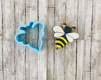 May include: A blue plastic cookie cutter shaped like a bee, next to a decorated sugar cookie shaped like a bee with yellow and black stripes and white wings.
