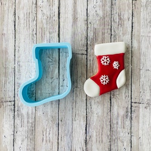May include: A blue plastic Christmas stocking cookie cutter next to a decorated Christmas stocking cookie. The cookie is red with white icing snowflakes.