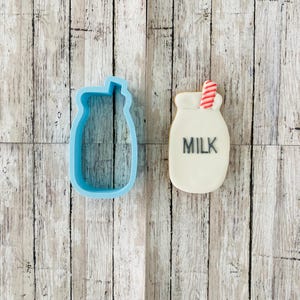 May include: A blue plastic cookie cutter shaped like a milk jug with a straw, next to a white iced cookie decorated with red and white stripes and the word "MILK" in black icing.