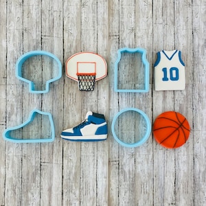 Puede incluir: Un conjunto de cortadores de galletas azules y blancos con forma de artículos con temática de baloncesto, que incluyen una pelota de baloncesto, una camiseta, zapatillas deportivas y un aro.