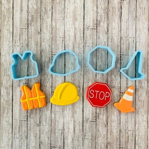 May include: A set of nine blue plastic cookie cutters in various shapes, including a vest, hard hat, stop sign, traffic cone, and octagon. The cutters are on a wood background.