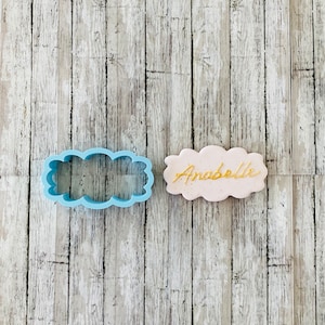May include: A blue plastic cookie cutter shaped like a cloud with a pink cloud cookie with the name "Anabelle" written in gold icing.