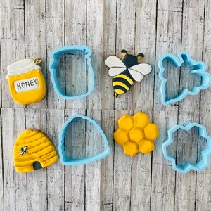 May include: A set of nine blue cookie cutters in various shapes, including a honey pot, bee, honeycomb, and beehive. The cutters are arranged on a wooden surface.