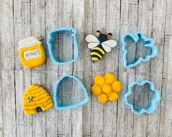 May include: A set of nine blue cookie cutters in various shapes, including a honey pot, bee, honeycomb, and beehive. The cutters are arranged on a wooden surface.
