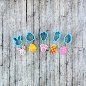 May include: A set of blue plastic cookie cutters shaped like a bunny, chick, paw print, carrot, egg, and a flower. There are also five decorated cookies, including a bunny, chick, paw print, carrot, and egg.