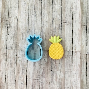 May include: A blue plastic pineapple cookie cutter and a yellow and green sugar cookie shaped like a pineapple.