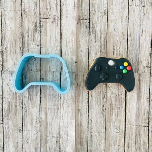 Może przedstawiać: Jasnoniebieska plastikowa foremka do ciastek w kształcie kontrolera do gier wideo leży obok ozdobionego ciastka. Ciastko jest czarne z kolorowymi przyciskami i białym środkowym przyciskiem. Tło to jasne usłojenie drewna.