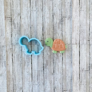 May include: A light blue plastic turtle-shaped cookie cutter next to a decorated turtle cookie. The cookie has a green head and feet, a brown shell, and a smiling face. The background is a light wood grain.
