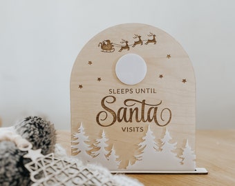 May include: Wooden Christmas countdown sign with the text "Sleeps Until Santa Visits". The sign features a laser-cut design of a sleigh with reindeer, stars, and a forest scene. A wooden Christmas tree ornament is also visible.