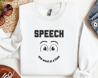 Puede incluir: Sudadera blanca con texto negro que dice "SPEECH one word at a time". El texto está en negrita, en una fuente de bloque. Debajo del texto hay una cara de dibujos animados con ojos grandes.
