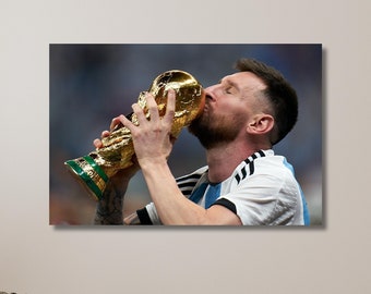 Puede incluir: Un hombre con una camiseta de fútbol blanca y azul besa el trofeo de la Copa Mundial de la FIFA. El trofeo es de oro y tiene detalles intrincados. El hombre sonríe y tiene barba.