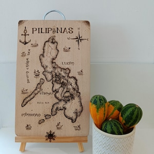 May include: A wooden map of the Philippines with the words "PILIPINAS" and "LUZON" burned into the wood. The map is on a small easel and there are gourds in a white pot with a chevron pattern next to it.