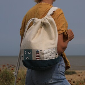 May include: A person wearing a yellow shirt and blue jeans is walking on a beach with a white drawstring backpack with a blue, green, and red lighthouse design.