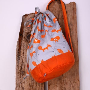 Può includere: Uno zaino a cordoncino grigio e arancione con una stampa di volpe. Lo zaino ha una lunga tracolla ed è appeso a una superficie di legno.