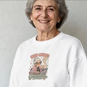 May include: A white crewneck sweatshirt featuring a graphic illustration of an older person with gray hair wearing glasses, sitting at a laptop with a cup of coffee and yarn, with the text VIBE-CODING GRANDMA in a retro, multi-colored font style.