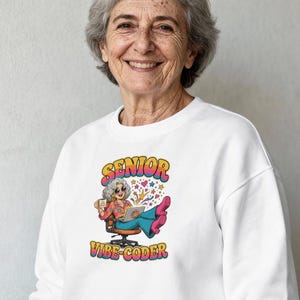 May include: A white crewneck sweatshirt featuring a graphic of a person with gray hair wearing sunglasses, a floral shirt, and blue pants, sitting in an office chair with a laptop. The text SENIOR VIBE-CODER is printed in colorful, retro-style lettering.