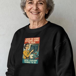 May include: A black crewneck sweatshirt featuring a graphic print of a woman in a yellow cardigan pointing at a chalkboard labeled Robotics 101. The text reads: SPENT 30 YEARS EXPLAINING THINGS TO MEN. NOW I EXPLAIN THINGS TO AI. A small robot sits nearby.