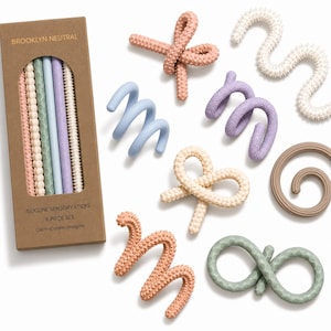 May include: A Brooklyn Neutral 8-piece silicone sensory stick set in a brown box with a window. The set includes various textured, flexible shapes in muted colors like peach, sage green, lavender, blue, cream, and tan, featuring spiral, wavy, and knotted designs.