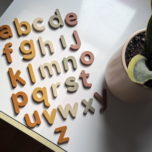 May include: A set of lowercase alphabet letters made of wood, arranged in rows. The letters feature a muted color palette of terracotta, mustard yellow, beige, and sage green. Each letter is cut in a rounded, sans-serif font style.