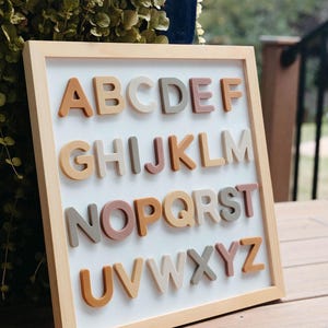May include: A framed wooden sign with the alphabet in colorful letters. The letters are made of a soft, textured material and are arranged in alphabetical order. The frame is a light wood color and the background is white.