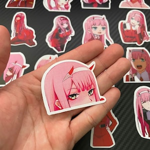 May include: A set of pink and red anime character stickers featuring a girl with pink hair and red eyes. The stickers are on a black background with a carbon fiber pattern.
