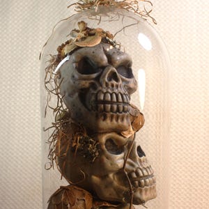 May include: A decorative glass cloche with a stack of three detailed skull sculptures. The skulls are a muted gray color, with dried leaves and straw-like material inside. The cloche sits on a glass pedestal.