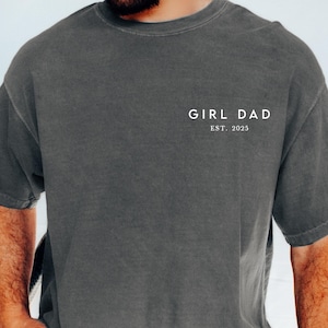 May include: Dark gray t-shirt with the text "GIRL DAD EST. 2025" in white. The shirt is short-sleeved and has a relaxed fit. The shirt is made of a soft material.