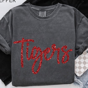 May include: A dark gray sweatshirt with the word "Tigers" in red glitter lettering. The word "Pepper" is in the top left corner. The sweatshirt is folded, with a phone case and jeans visible in the background.