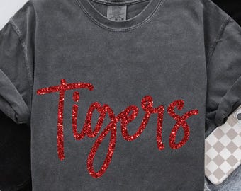 May include: A charcoal grey short-sleeved t-shirt with a crew neckline and a vintage-washed finish. The front features the word Tigers written in a cursive, red glitter script. The text PEPPER is printed in the upper left corner of the image.