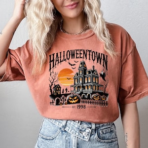 K&ouml;nnte beinhalten: Ein orangefarbenes T-Shirt mit einem grafischen Aufdruck eines Spukhauses, eines Sonnenuntergangs und dem Text "HALLOWEENTOWN EST. 1998".