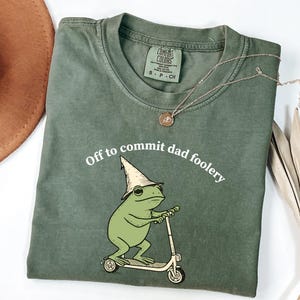 May include: Olive green t-shirt with the phrase "Off to commit dad foolery" and a cartoon frog riding a scooter. The frog wears a pointed hat. A gold necklace and a brown hat are also visible.