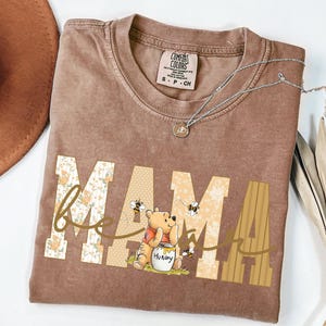 May include: A brown Comfort Colors t-shirt featuring the word "MAMA" in a floral and striped design, with a Winnie the Pooh graphic. The shirt has a crew neck and short sleeves. A silver necklace with a pendant is also visible.