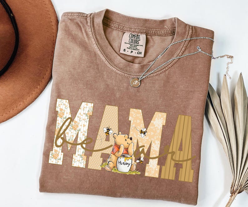 May include: A brown Comfort Colors t-shirt featuring the word "MAMA" in a floral and striped design, with a Winnie the Pooh graphic. The shirt has a crew neck and short sleeves. A silver necklace with a pendant is also visible.