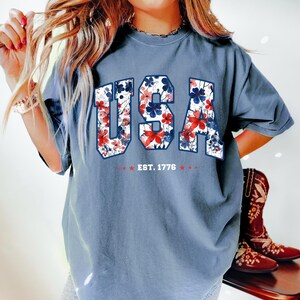 May include: A blue t-shirt with the word "USA" in a floral design of red, white, and blue. The text "EST. 1776" is below the word "USA". The shirt is a comfortable fit.