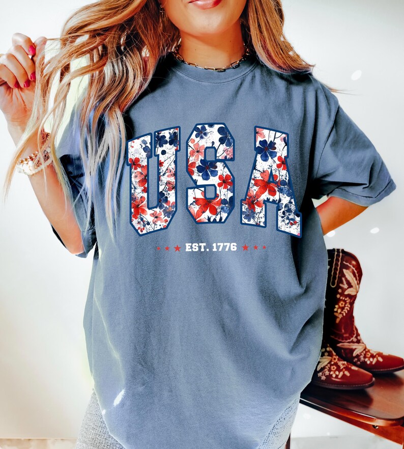 May include: A blue t-shirt with the word "USA" in a floral design of red, white, and blue. The text "EST. 1776" is below the word "USA". The shirt is a comfortable fit.