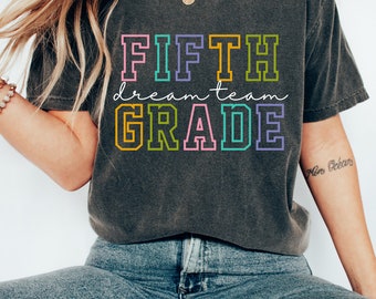 Puede incluir: Una camiseta gris oscuro con el texto "FIFTH dream team GRADE" en colores arcoíris. El texto está contorneado en negro.