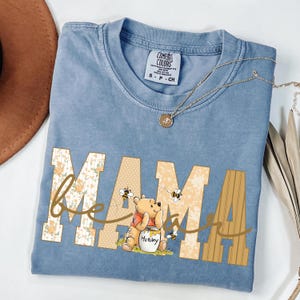 May include: A light blue Comfort Colors t-shirt with the word "MAMA" in a floral and honey-themed design. The shirt features a Winnie the Pooh graphic with bees and the word "bee" in cursive.