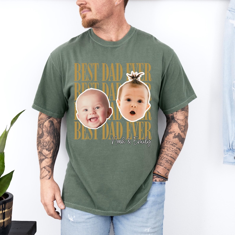 Może przedstawiać: Zielona koszulka w kolorze oliwkowym ze złotym napisem "BEST DAD EVER". Koszulka przedstawia dwie twarze dziecka oraz imiona "Noah & Emily". Koszulka ma swobodny styl.