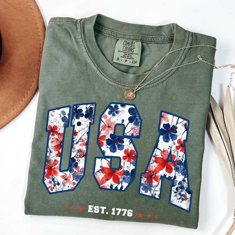 May include: Olive green t-shirt with the word "USA" in a floral design, outlined in blue. The shirt also features the text "EST. 1776" in red. A gold necklace and a brown hat are also visible.