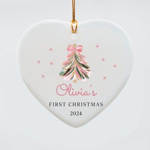 May include: White heart-shaped ceramic ornament with a pink and green watercolor Christmas tree with a pink bow. The ornament has the text "Olivia's First Christmas 2024" printed on it.