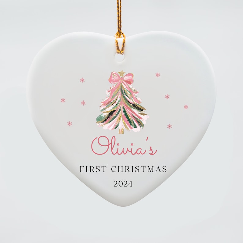 May include: White heart-shaped ceramic ornament with a pink and green watercolor Christmas tree with a pink bow. The ornament has the text "Olivia's First Christmas 2024" printed on it.