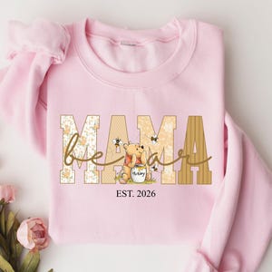 Op de afbeelding: Een lichtroze sweatshirt met het woord "MAMA" in een blokletterontwerp. Het woord "bear" is in cursief over de letters geschreven. Een Winnie de Poeh-afbeelding is te zien, samen met de tekst "EST. 2026".