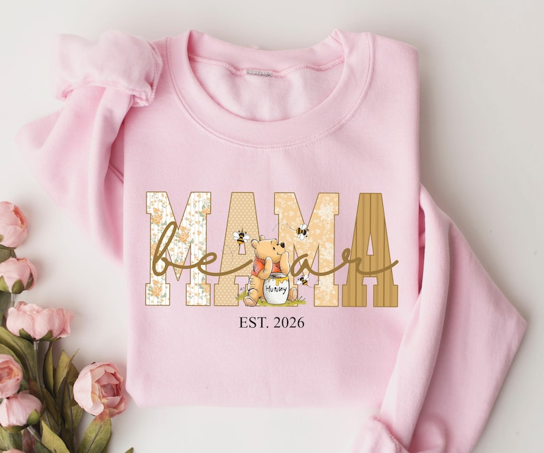 Op de afbeelding: Een lichtroze sweatshirt met het woord "MAMA" in een blokletterontwerp. Het woord "bear" is in cursief over de letters geschreven. Een Winnie de Poeh-afbeelding is te zien, samen met de tekst "EST. 2026".